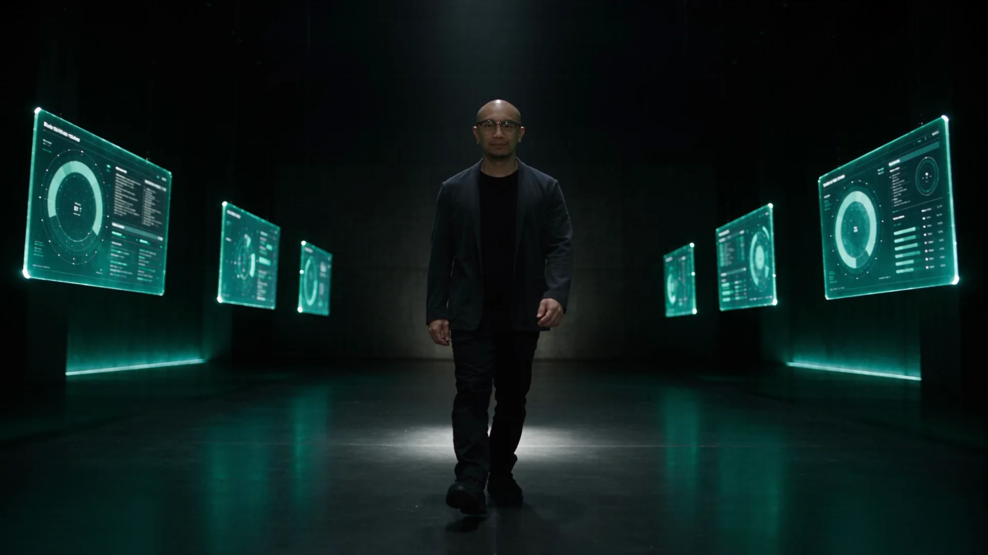 Nelson walking towards camera down a dark gallery of glowing dashboard screens